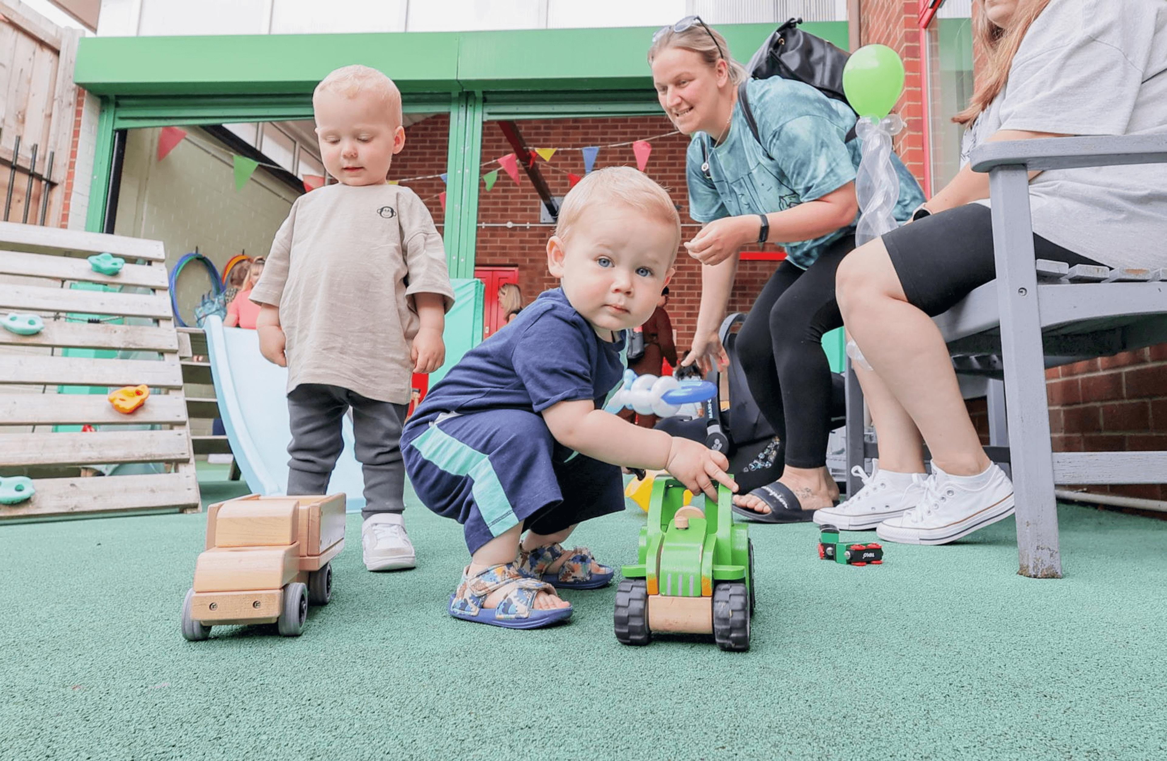 2 toddlers playing with toys in an outdoor play area while their parent/carer watches on